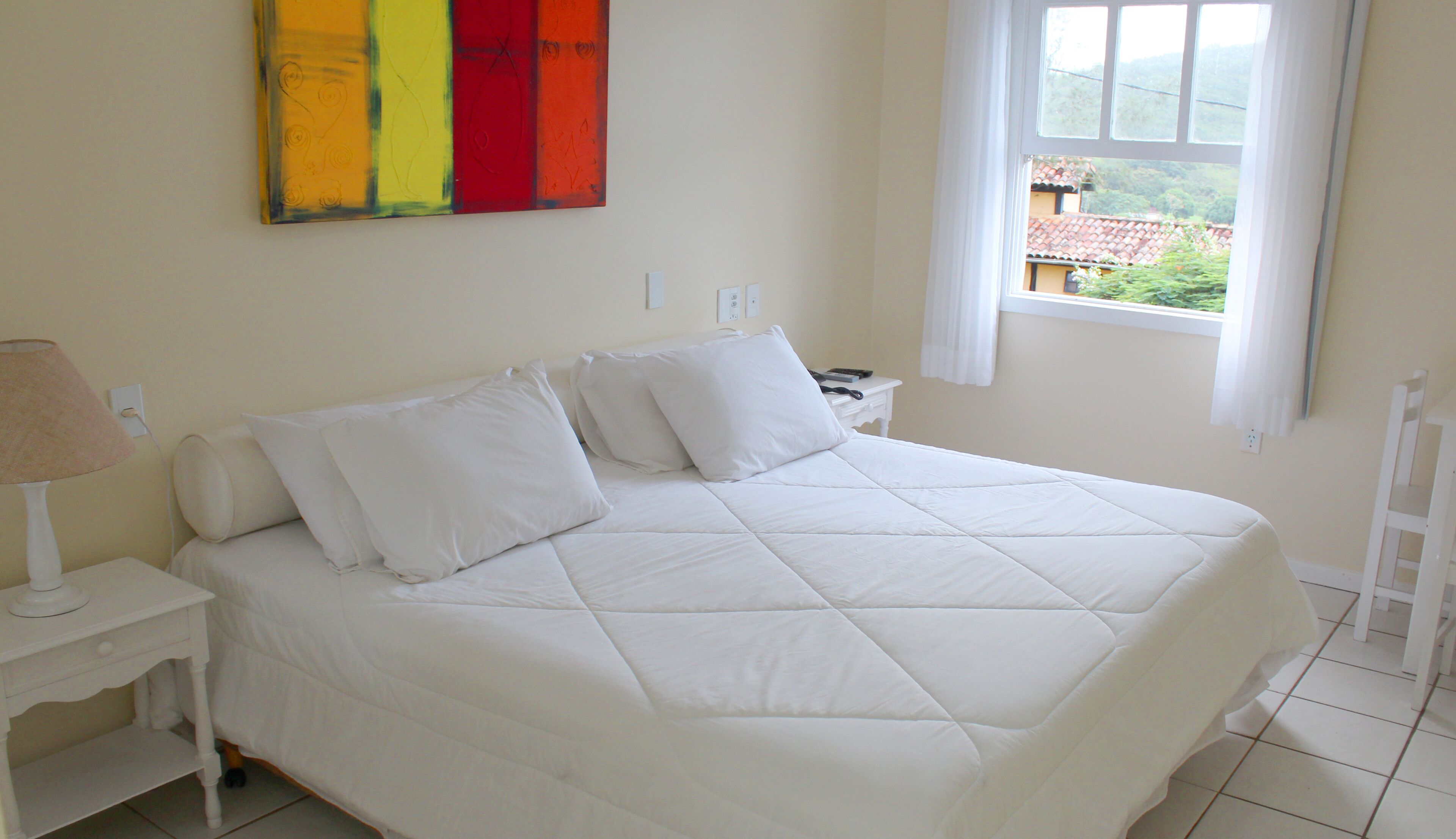 Un letto rifatto con lenzuola bianche in una stanza con una finestra, un comodino con una lampada e un quadro astratto dai colori vivaci appeso al muro.
