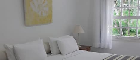 Una cama bien hecha con ropa blanca y un plaid a rayas, una lámpara de noche, una ventana con cortinas y un cuadro abstracto amarillo en la pared.