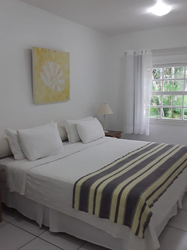 Una cama bien hecha con ropa blanca y un plaid a rayas, una lámpara de noche, una ventana con cortinas y un cuadro abstracto amarillo en la pared.