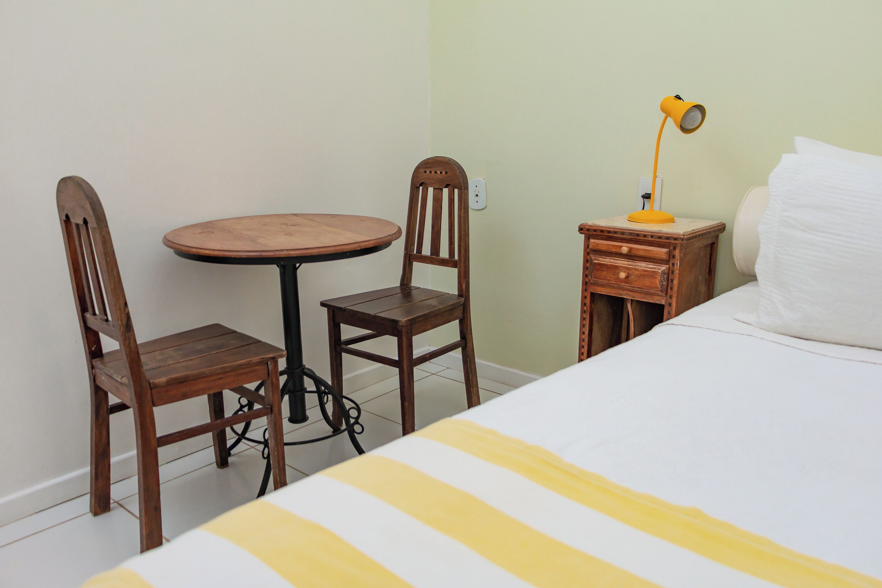A small room with a bed, a wooden table, two chairs, a nightstand with a lamp, and a power outlet on the wall.