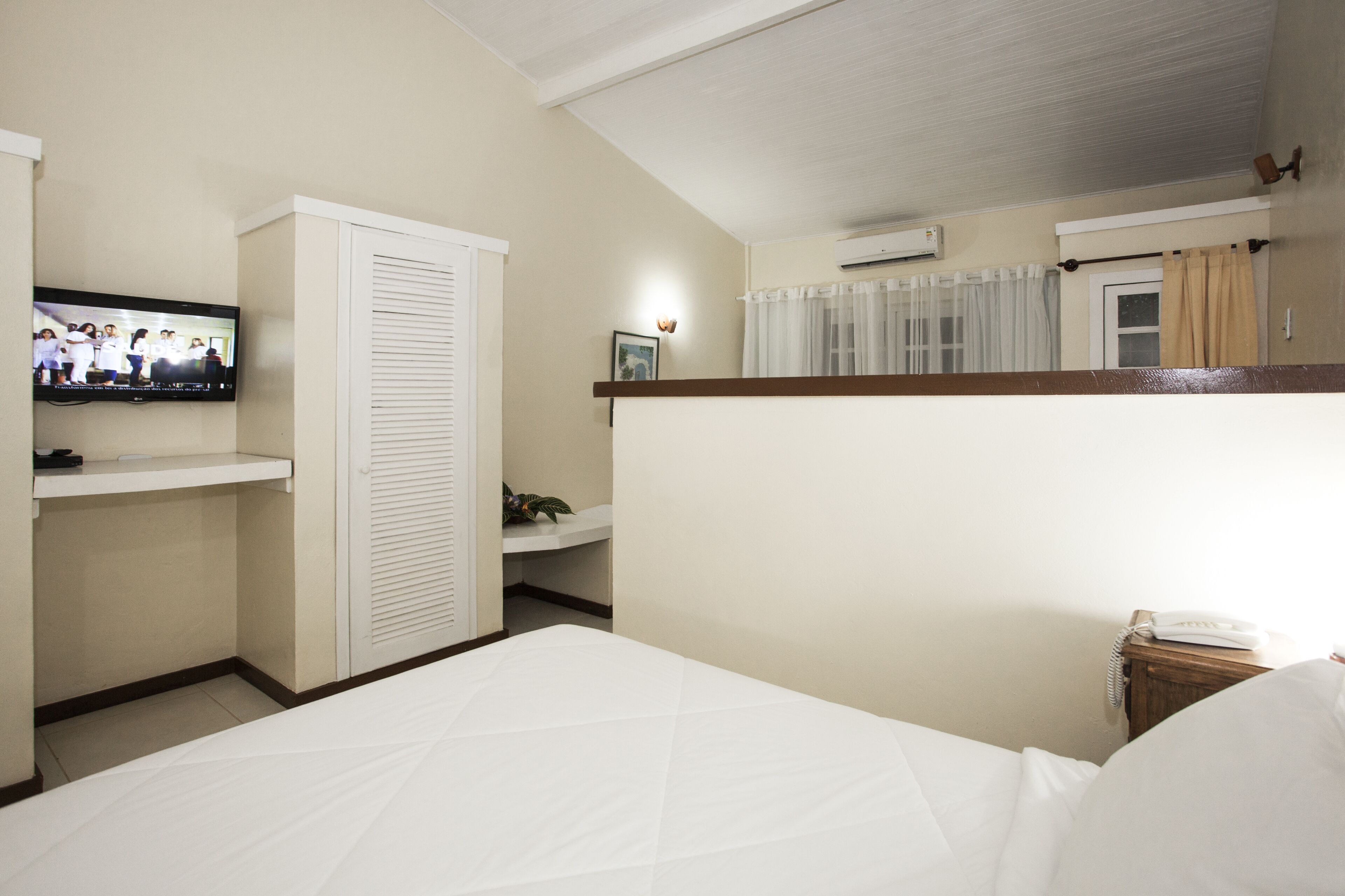 Une chambre d’hôtel avec un lit, un téléviseur fixé au mur, une armoire, une table de chevet avec une lampe et une petite plante.