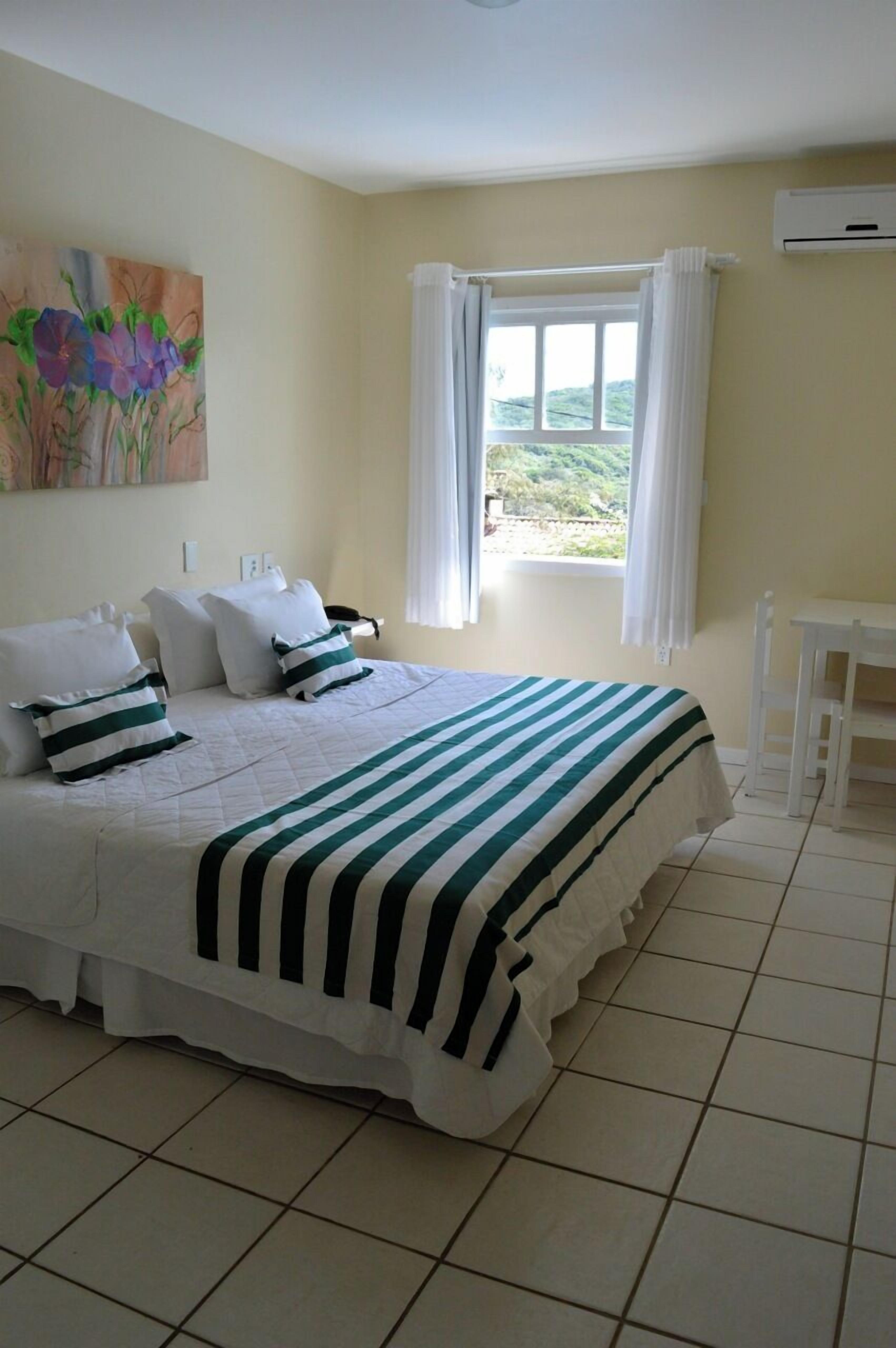 A neatly arranged bedroom with a large bed, white curtains, a small table, and a painting on the wall.