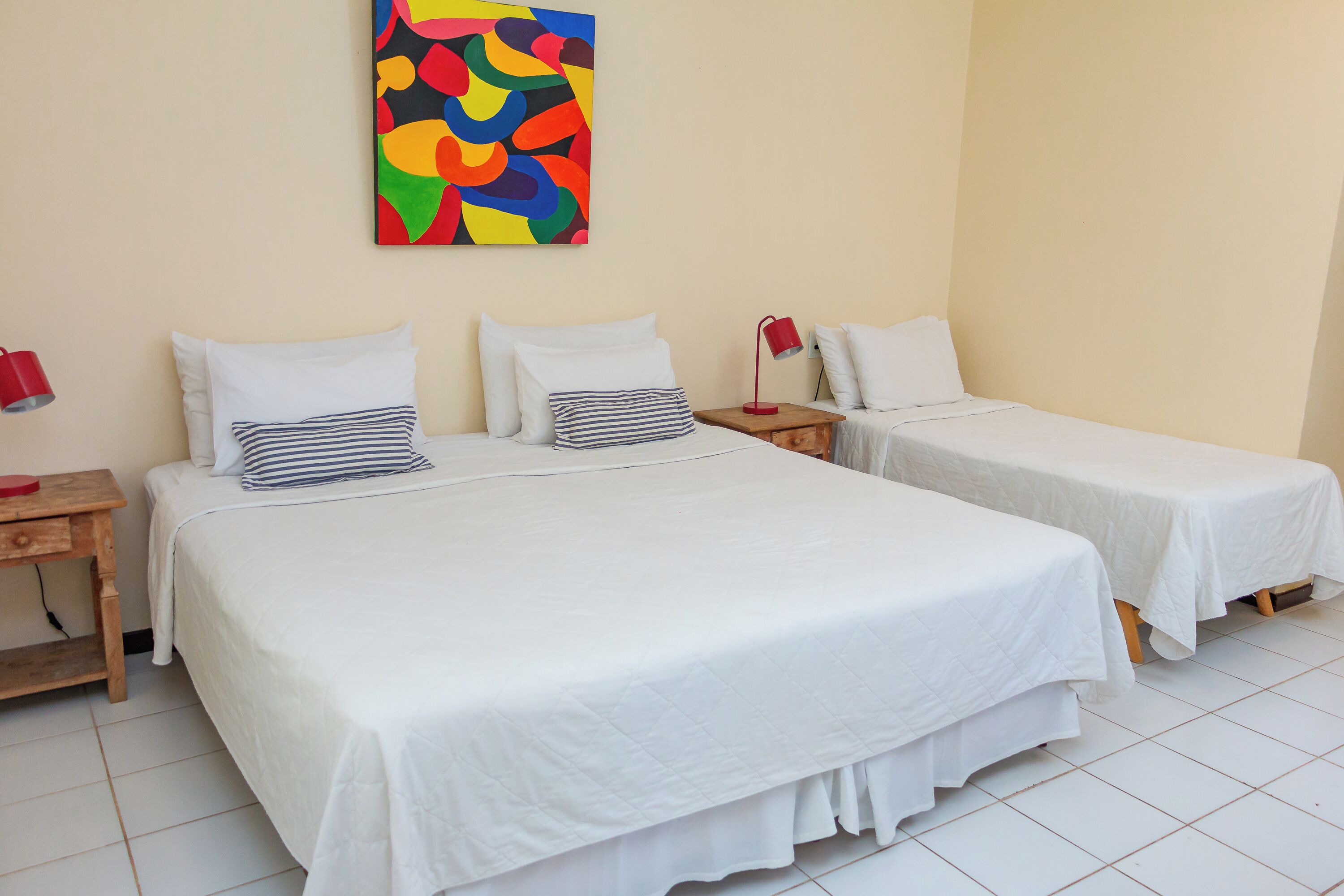 A hotel room with two single beds, a wooden nightstand with a red lamp, and a colorful abstract painting on the wall.