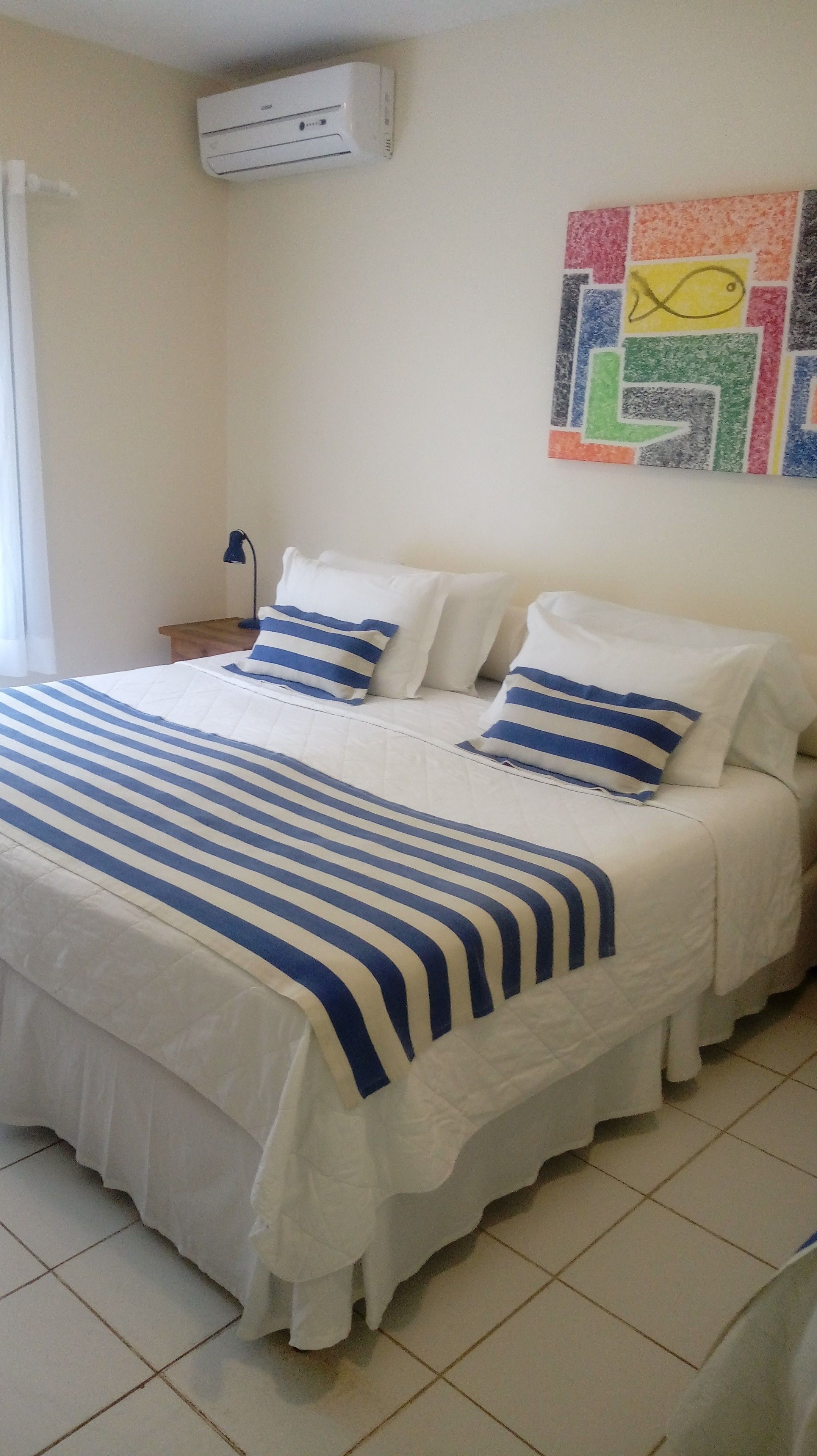 Camera da letto con un letto, cuscini, un comodino, una lampada, un condizionatore d'aria e un decoro vivace al muro.