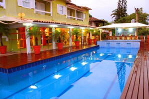 A swimming pool with a wooden deck and potted plants, surrounded by a building with multiple windows and a tiled roof. - Pousada Dos Reis (Búzios)