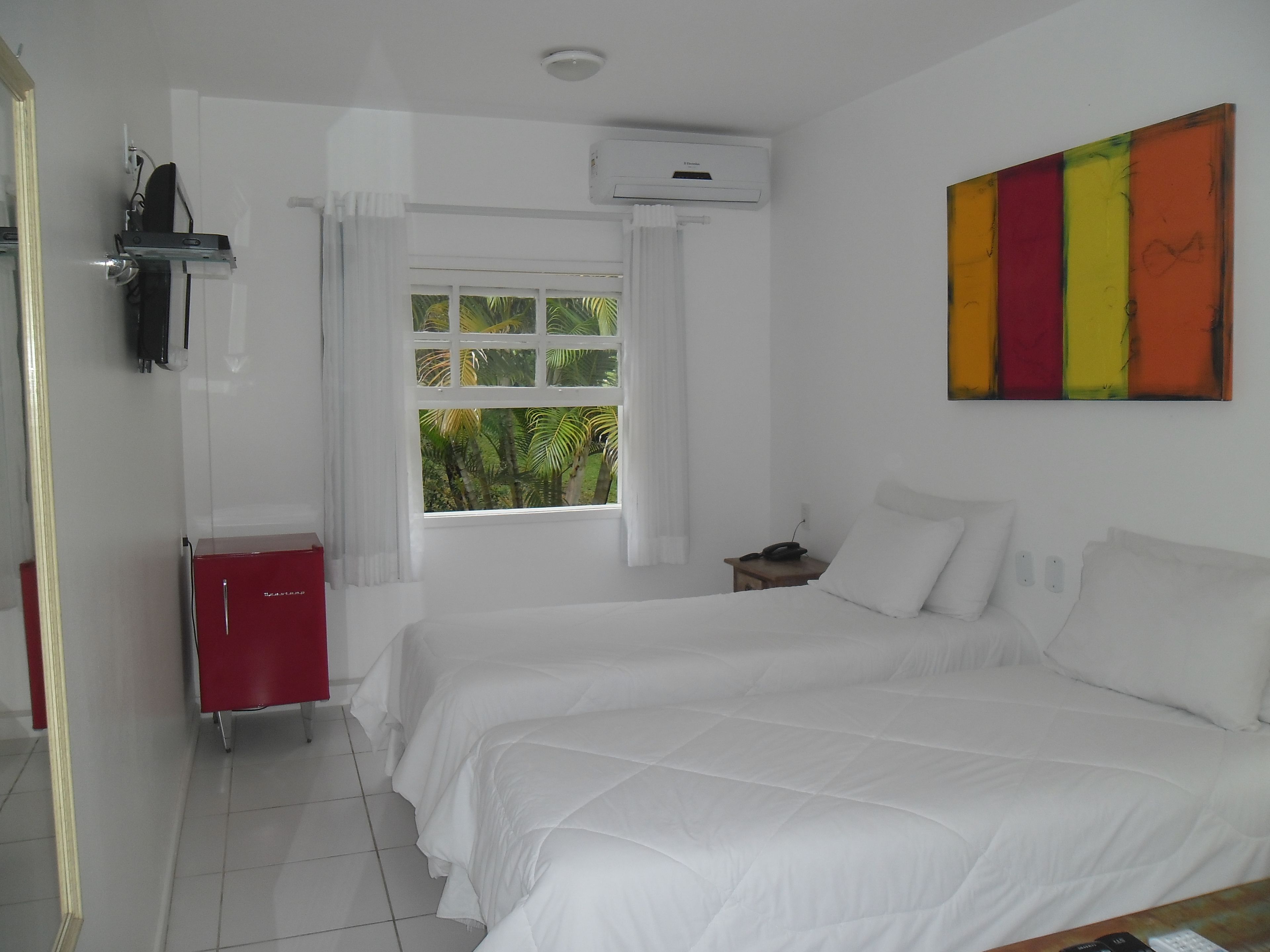 Una habitación de hotel con dos camas, un armario rojo, un ventanal con cortinas, un equipo de aire acondicionado y un cuadro de colores en la pared.