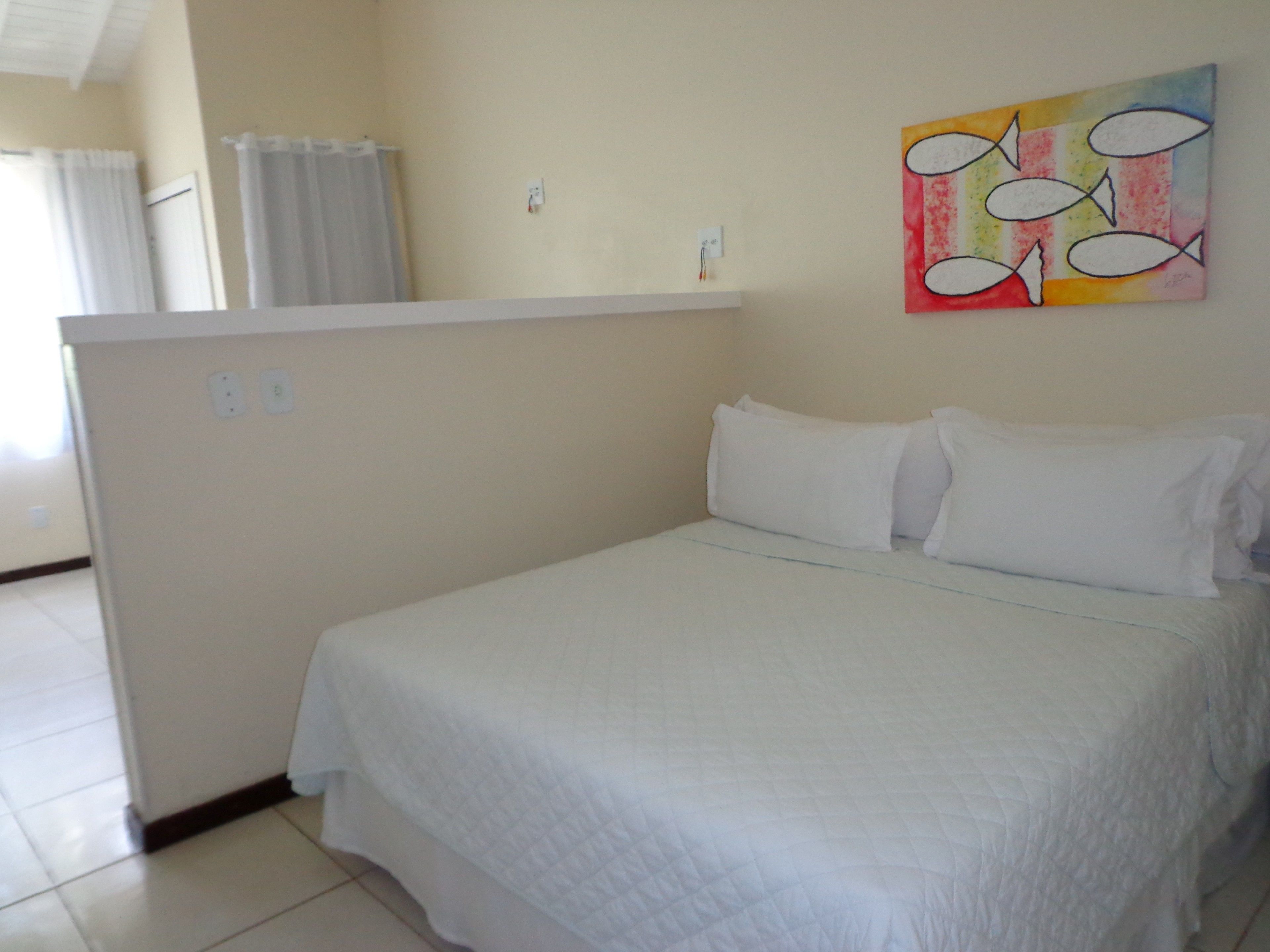 A bedroom with a bed, white curtains, a painting on the wall, and a small white table.