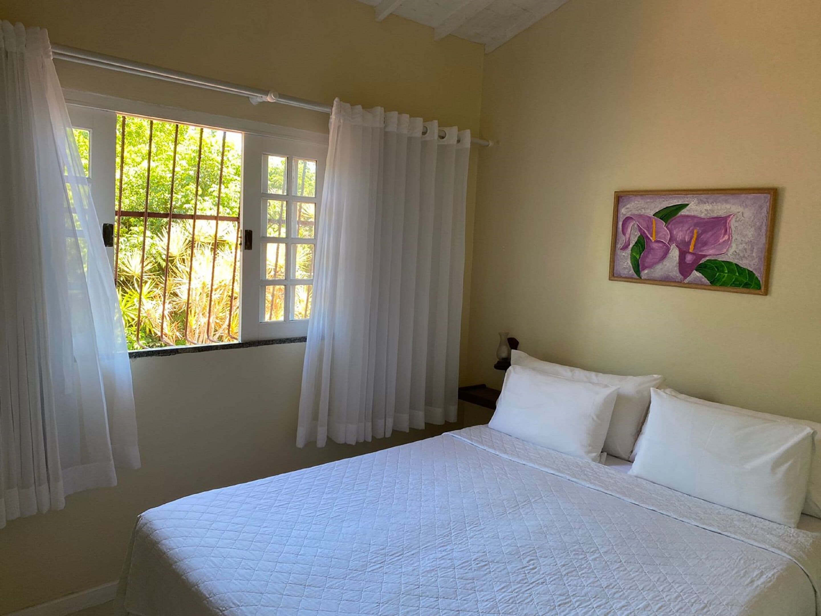 A bedroom with a large bed, white curtains, a window with a view of greenery, and a painting on the wall.
