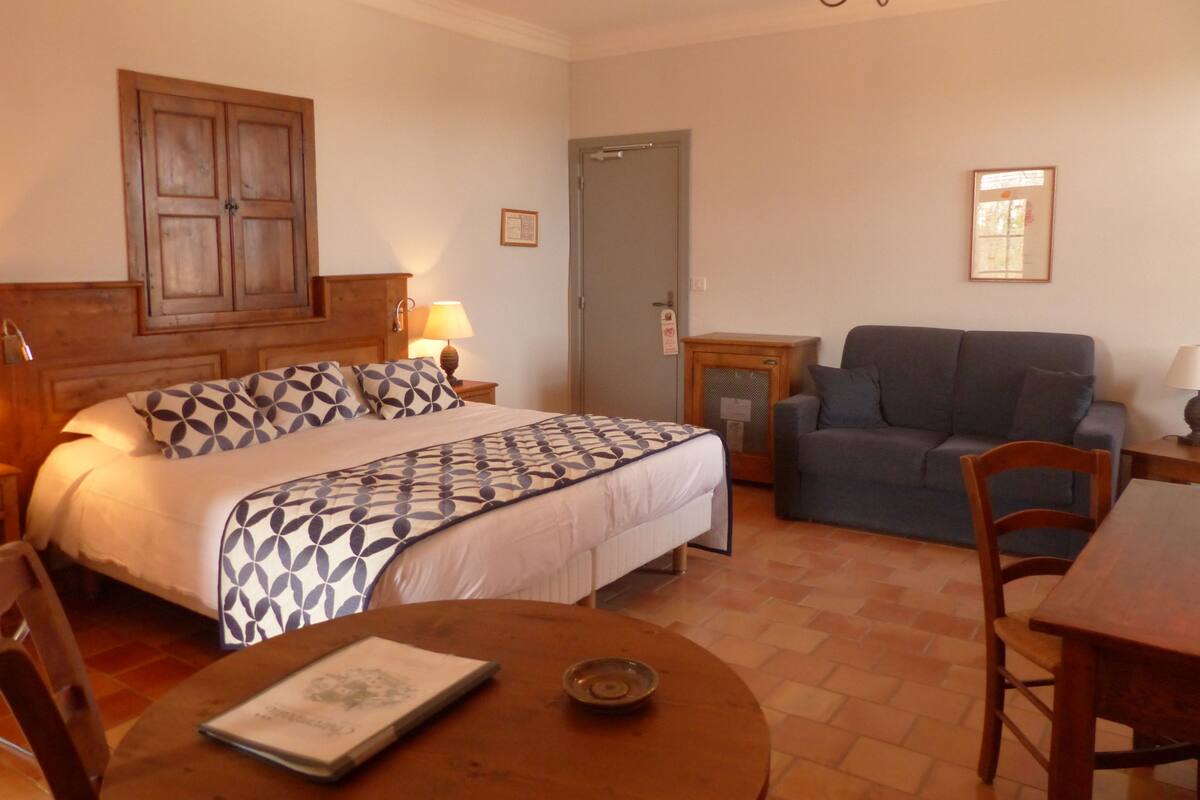 A bedroom with a bed, a wooden headboard, a bedside table with a lamp, a grey sofa, a wooden table with a book, and a door.