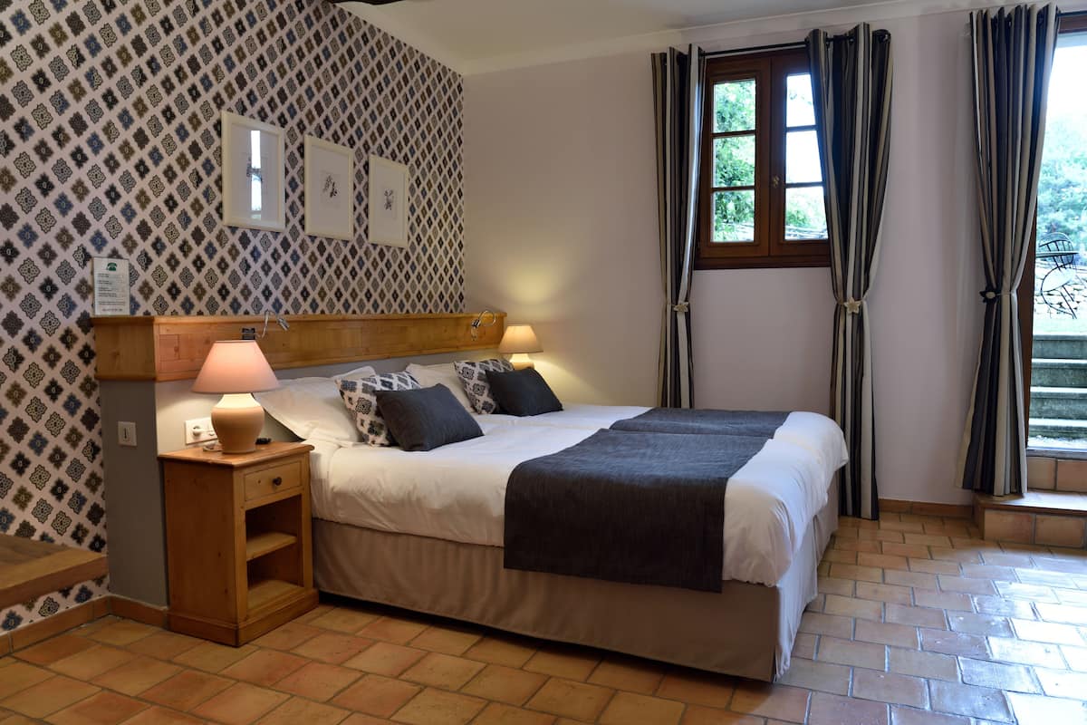 A double bed with a headboard, two bedside tables with lamps, patterned wallpaper, a window with curtains, and a tiled floor.