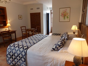 Una habitación de hotel con una cama grande, dos mesitas de noche con lámparas, un escritorio de madera con una silla, una pequeña mesa con sillas y una puerta que da a otra habitación.