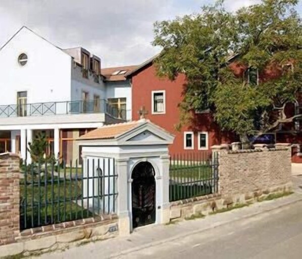 A two-story building with a white facade and a red section. There is a gated entrance with a small archway and a metal fence.