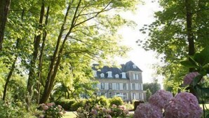 Un grand bâtiment historique à étages, doté d’une toiture remarquable et entouré d’une végétation luxuriante et de plantes à fleurs.