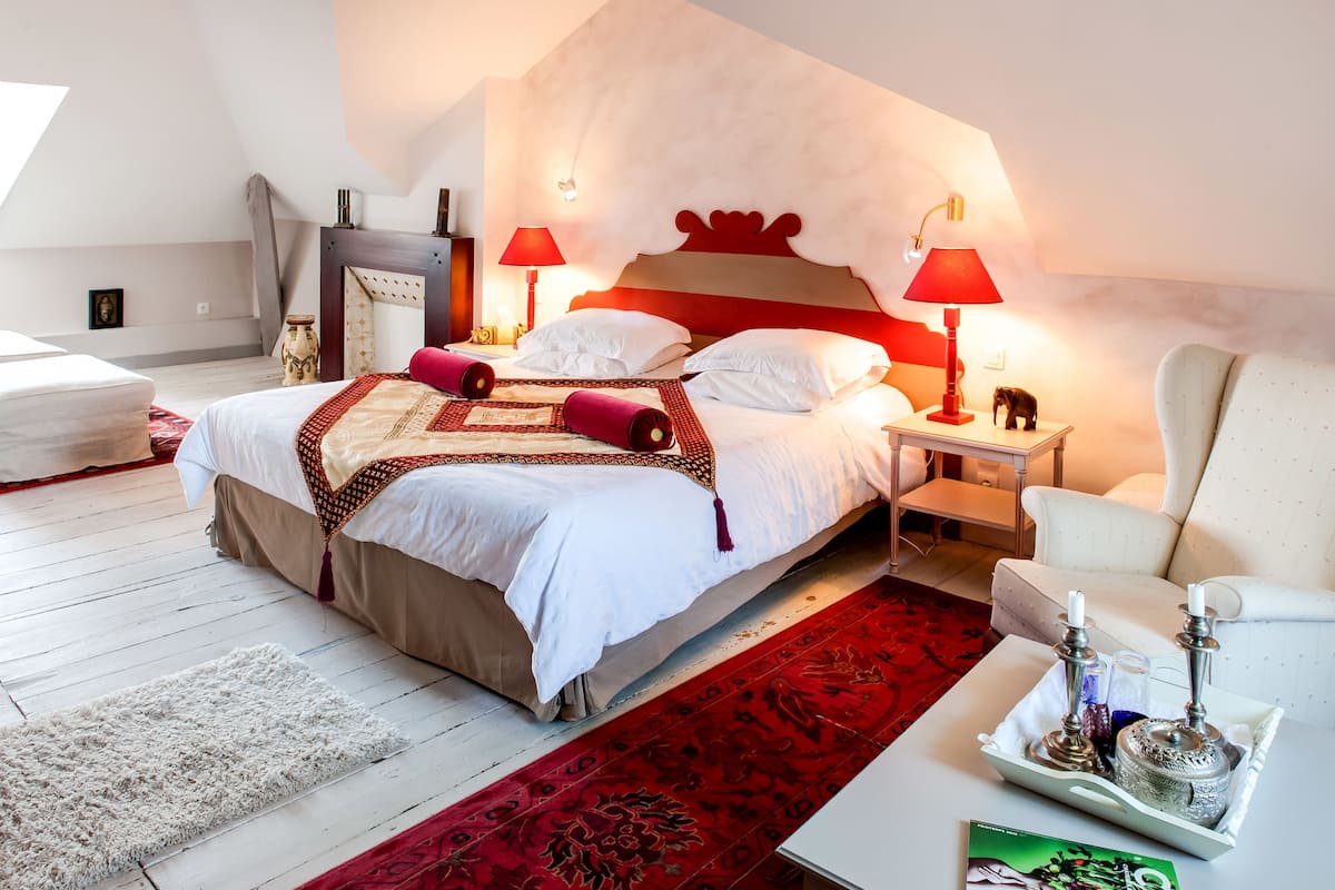 Une chambre d’hôtel moderne avec un grand lit, deux tables de chevet, un tapis aux motifs rouges et blancs, et un petit plateau décoratif avec des bougies.