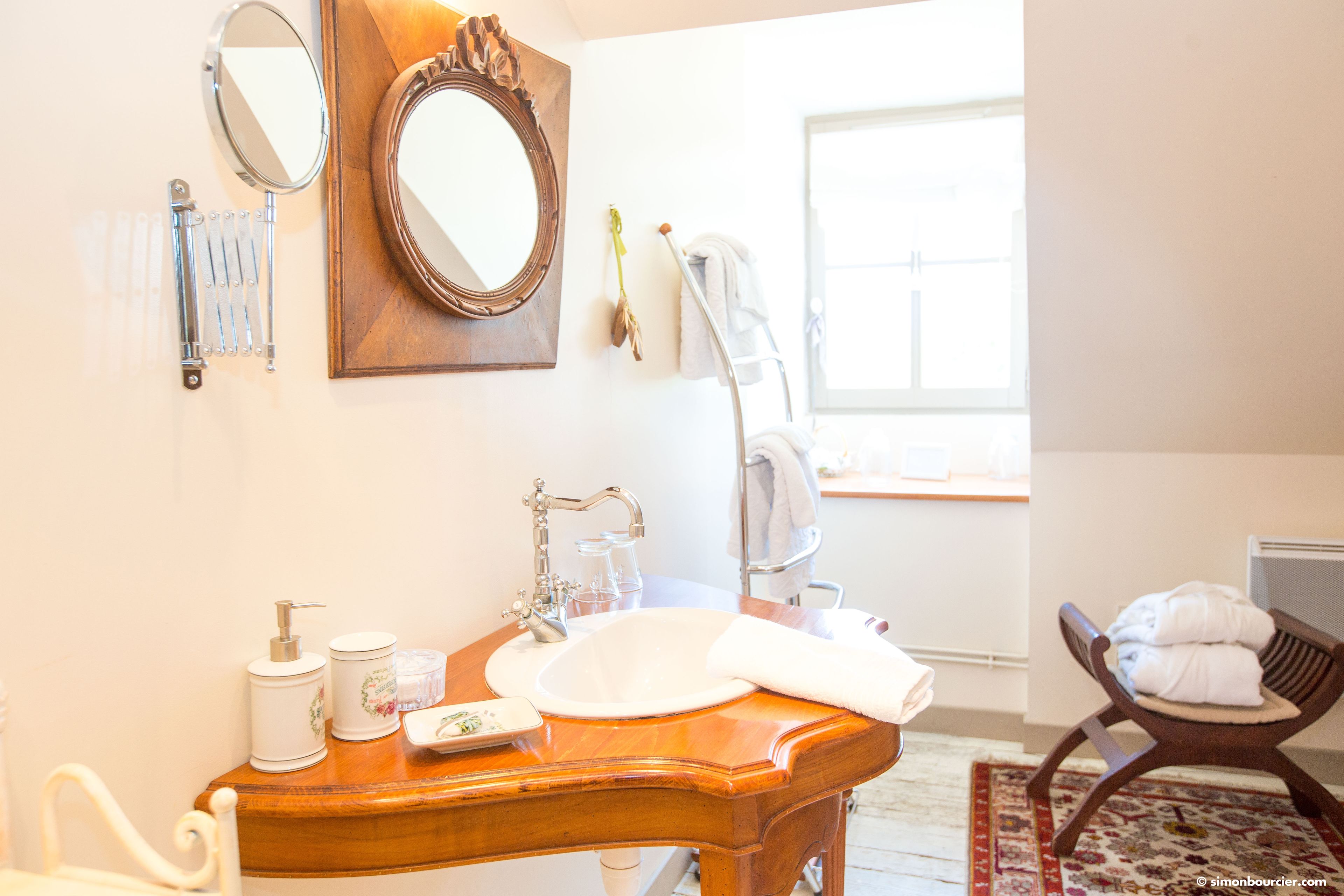 Un baño con tocador de madera, lavamanos, espejo, perchero para toallas y una silla.