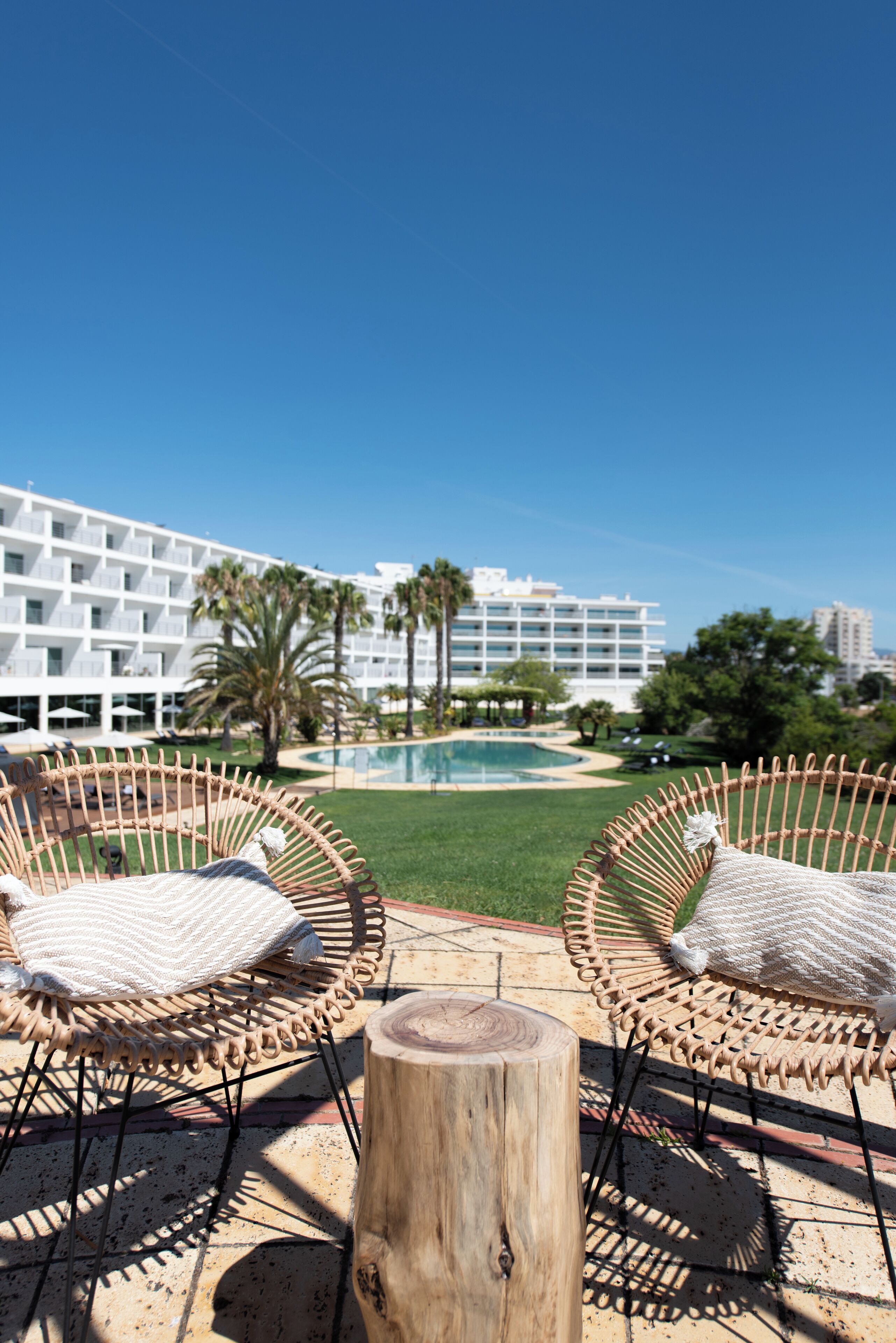A poolside area with wicker chairs and a wooden stump, overlooking a pool and a white building with balconies.