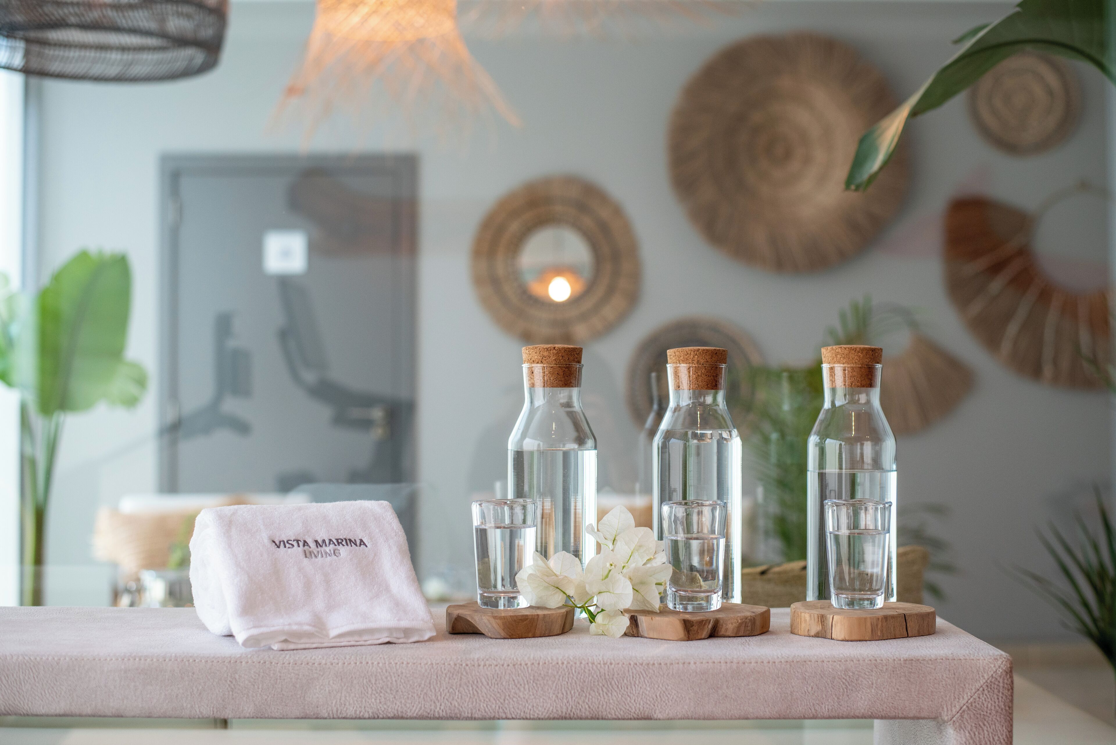 Ein ordentlich gedeckter Tisch mit vier Glasflaschen, einem weißen Handtuch und weißen Blumen auf einer rosafarbenen Oberfläche.