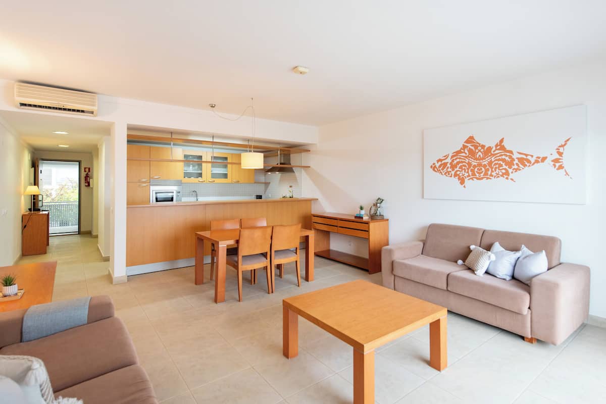 A modern living room with a wooden coffee table, a beige sofa, and a dining area with wooden chairs and a kitchen in the background.