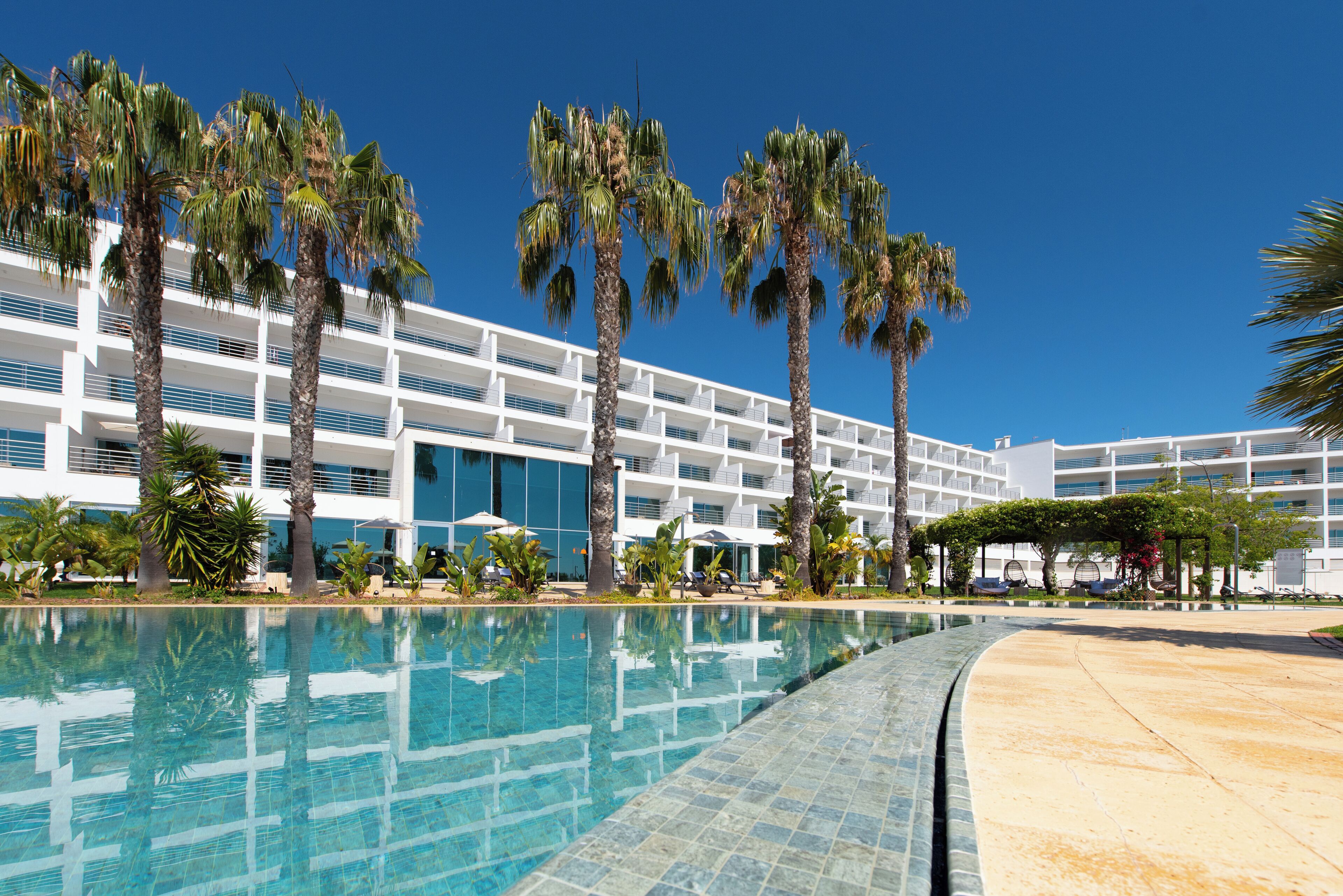 Una piscina amb aigua turquesa cristal·lina, envoltada de palmeres i un edifici d'hotel modern amb diversos pisos i balcons.