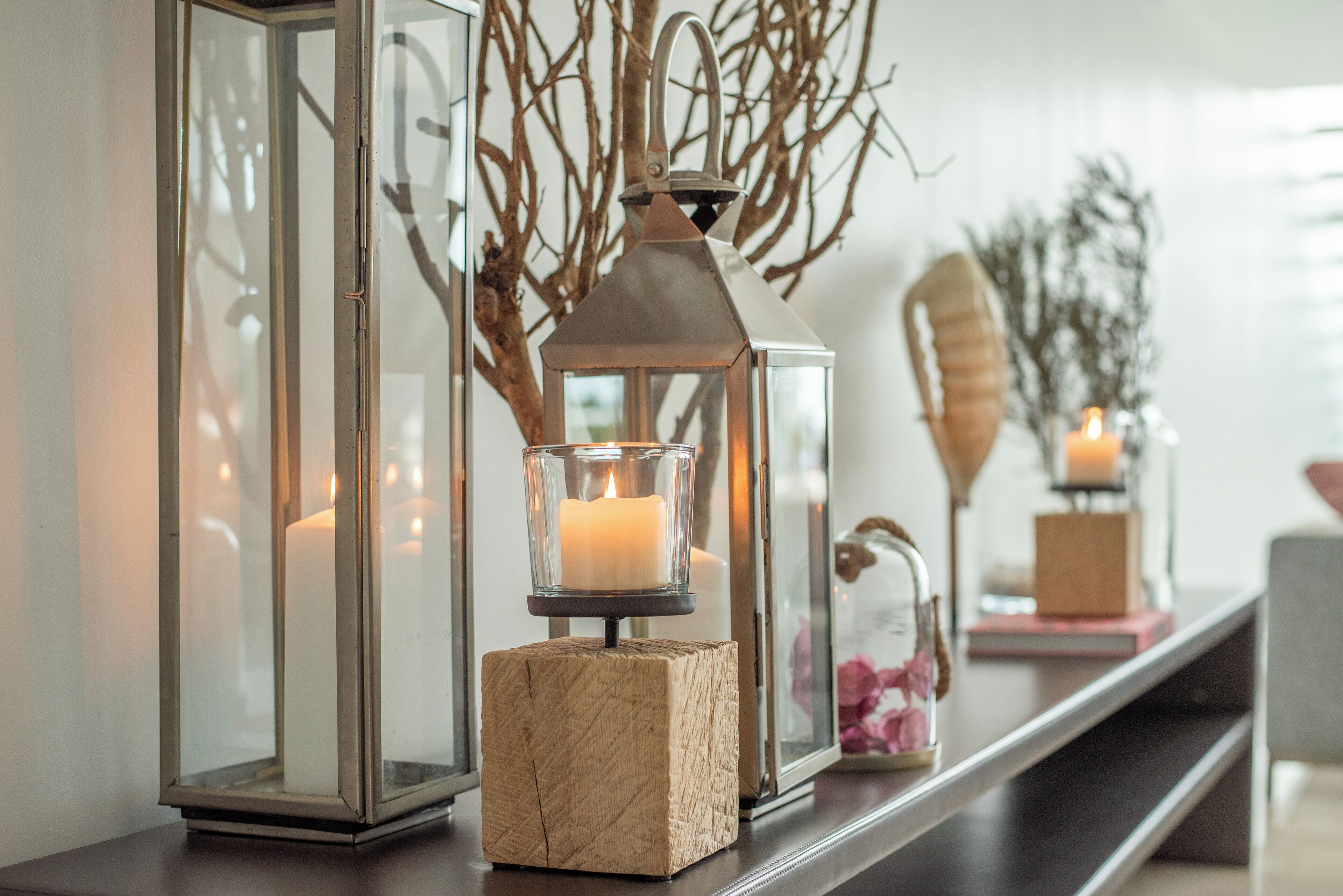 A row of glass lanterns with candles on a dark wooden surface, set against a white wall with decorative branches and a candle in the background.