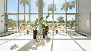 Un espacio interior amplio con ventanas grandes, plantas en macetas, sillas de mimbre y suelo de baldosas.