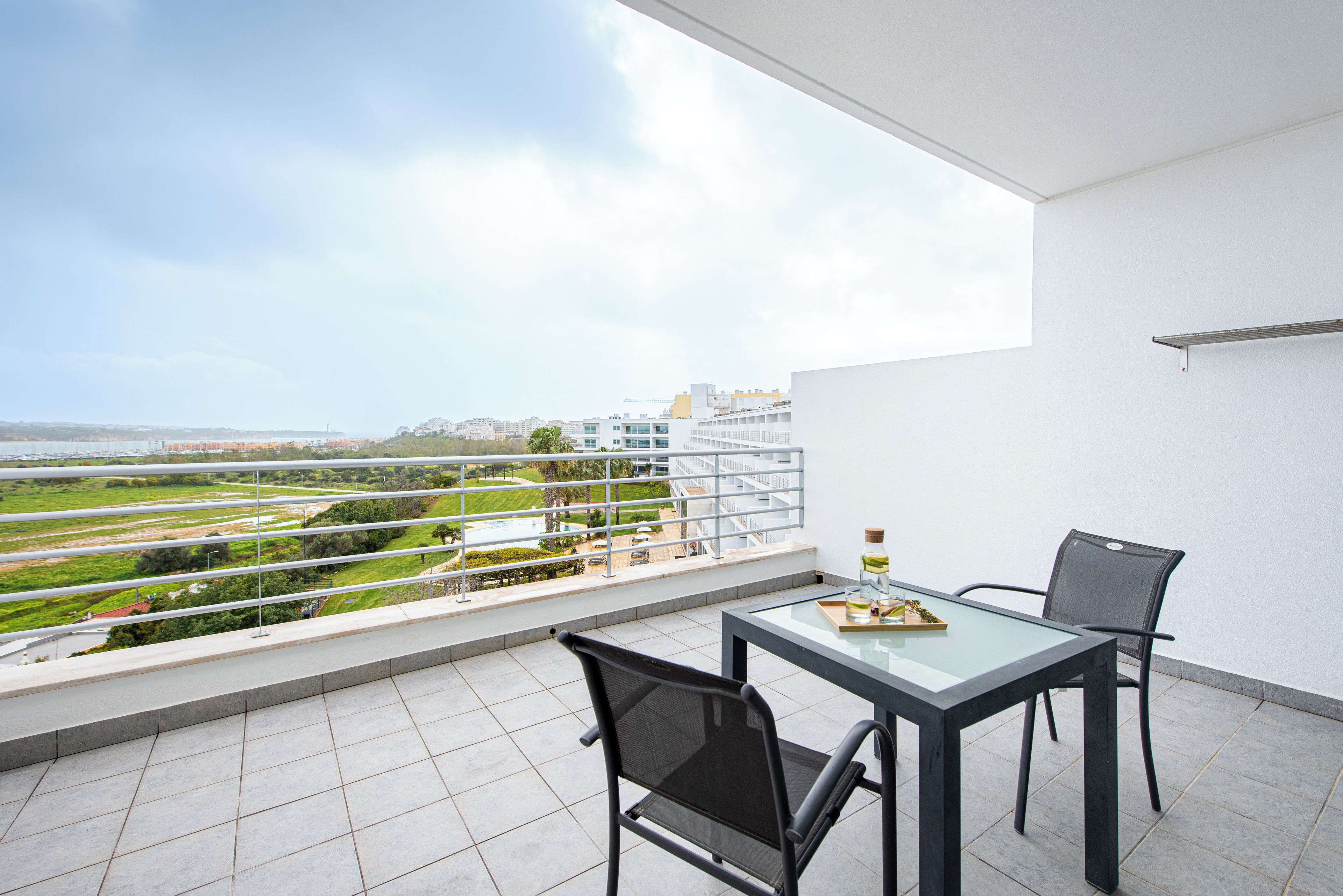 A balcony with a glass table and chairs, overlooking a cityscape and greenery.
