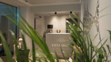 A reception area with a sign reading "VISTA MARINA SPA & WELLNESS," featuring a modern design with a reception desk, a wall-mounted lamp, and decorative elements like candles and plants.