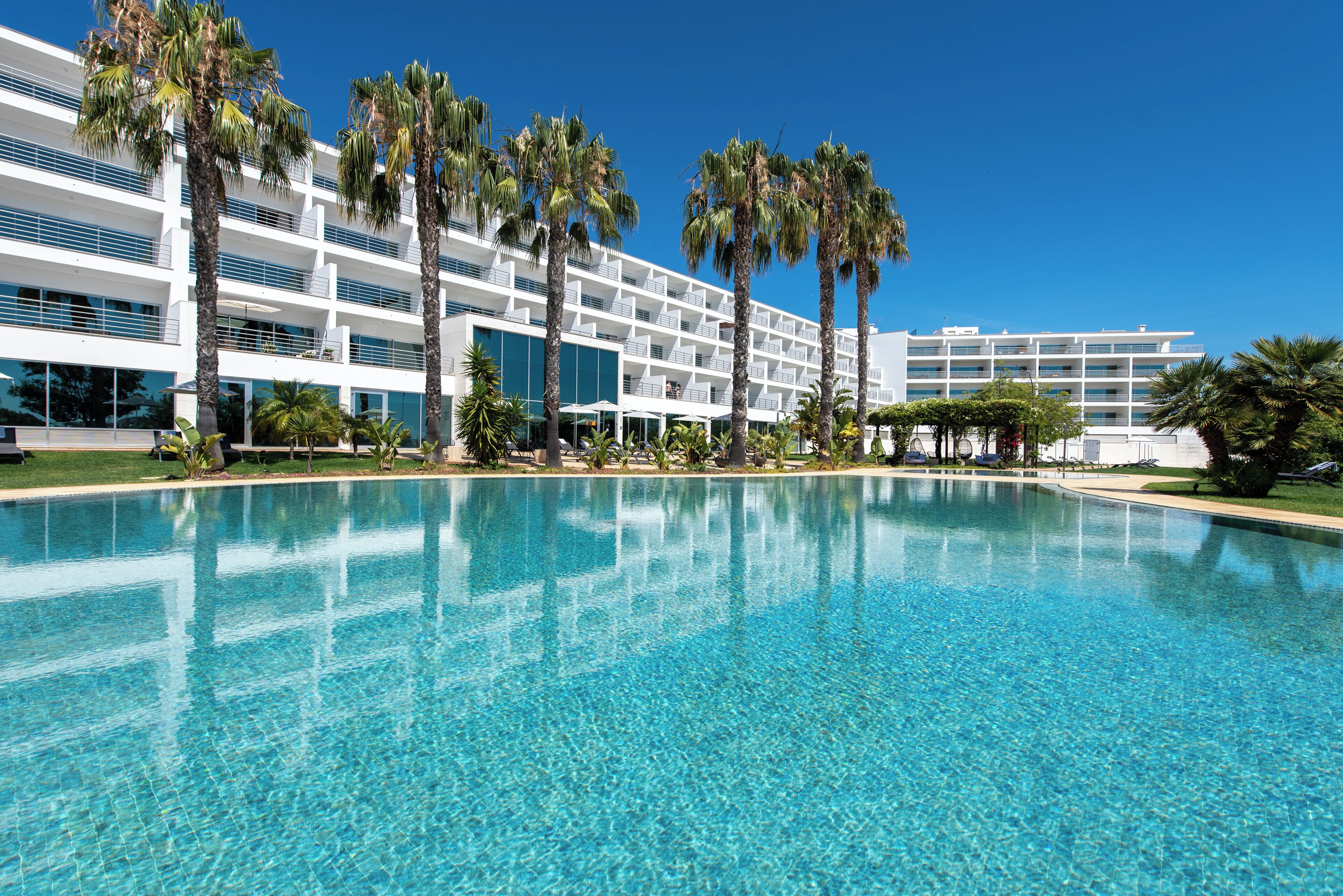 Un hotel moderno con una piscina grande, palmeras y un cielo azul despejado.