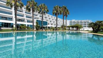 Un hotel moderno con una piscina grande, palmeras y un cielo azul despejado.
