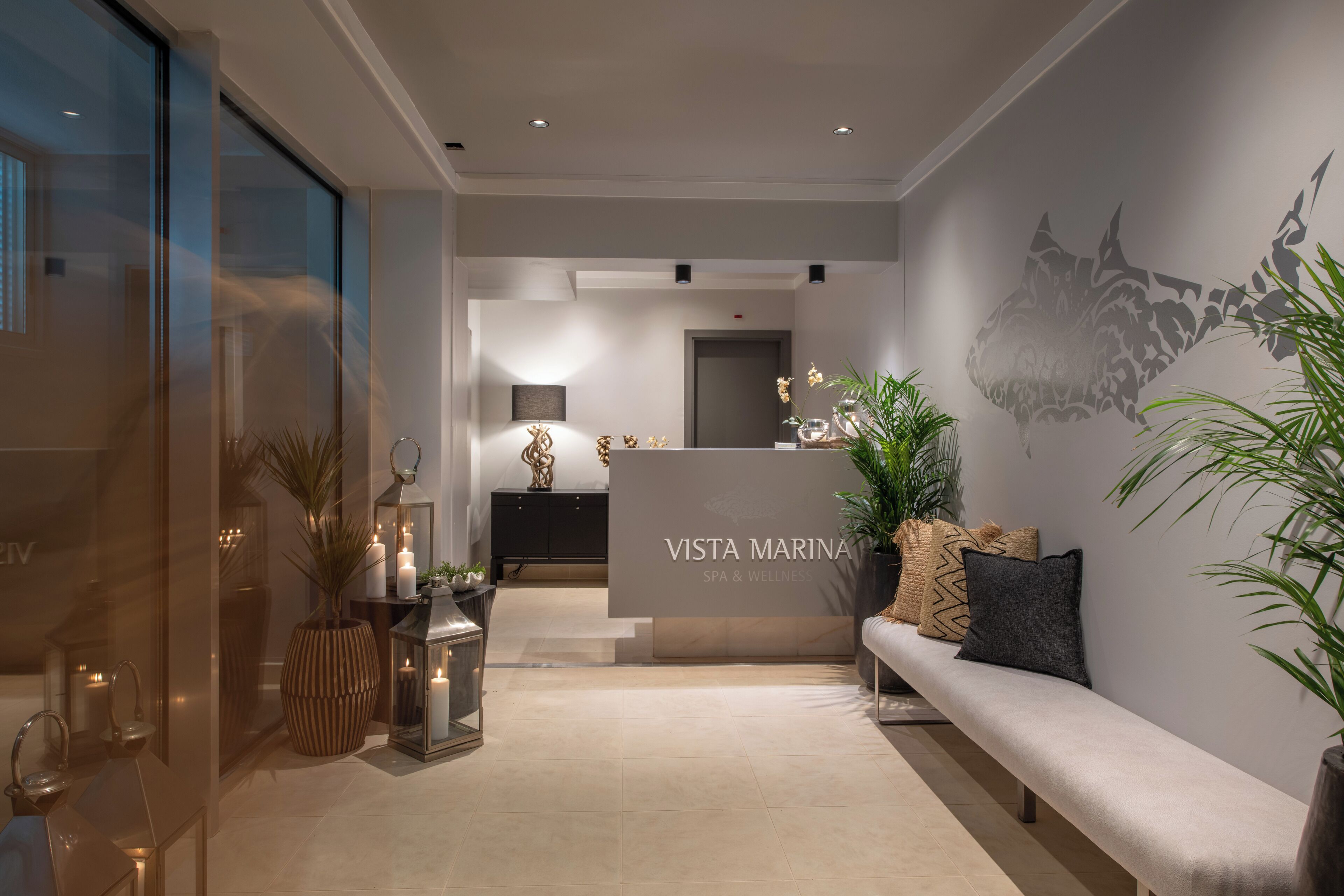 A modern reception area with a reception desk labeled "VISTA MARINA SPA & WELLNESS." There is a bench with decorative pillows, potted plants, and candles on the floor.