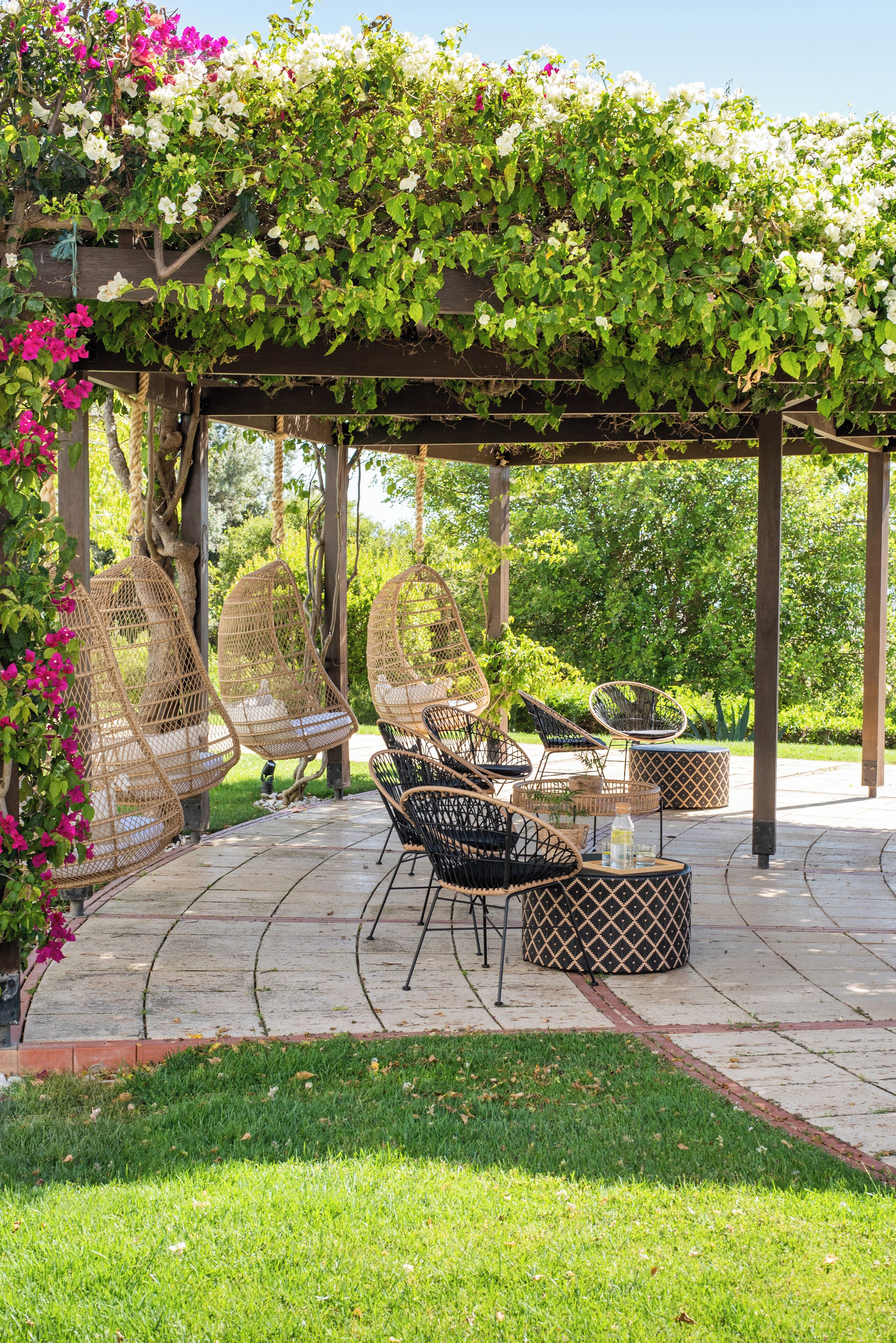 A covered outdoor seating area with wicker chairs and a table, surrounded by lush greenery and flowering plants.