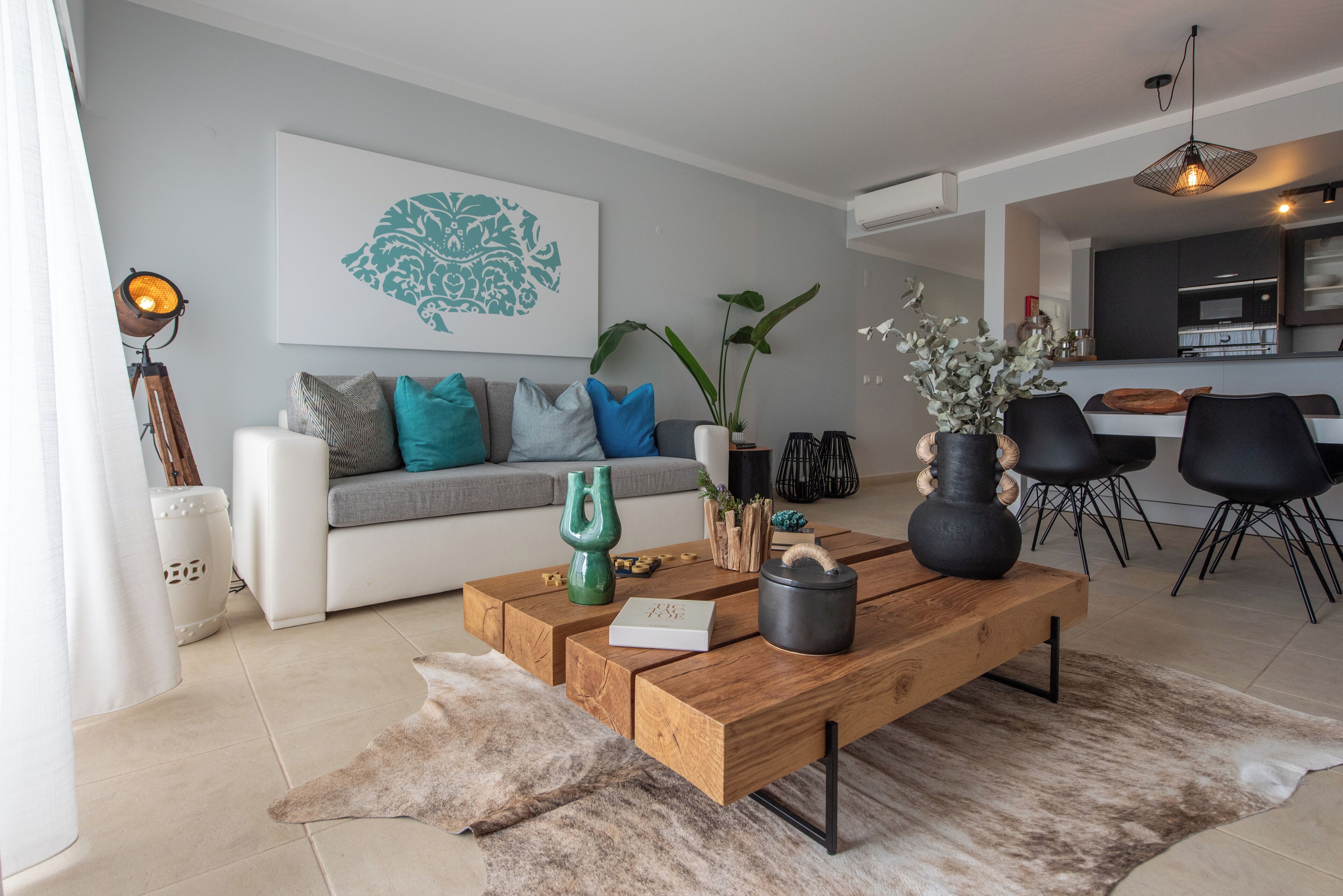 A modern living room with a wooden coffee table, a sofa with cushions, a potted plant, and a kitchen in the background.