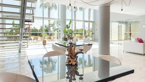 Un comedor amplio y moderno con una mesa de vidrio, sillas blancas y una pieza decorativa central. Grandes ventanales ofrecen vistas a palmeras y a un balcón.