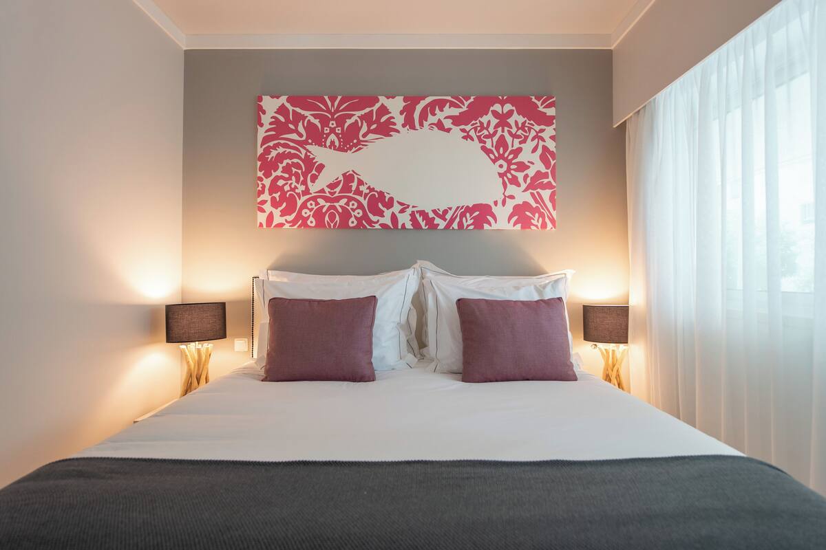 A neatly made bed with white and purple pillows, a large floral wall art piece above it, and two bedside lamps.