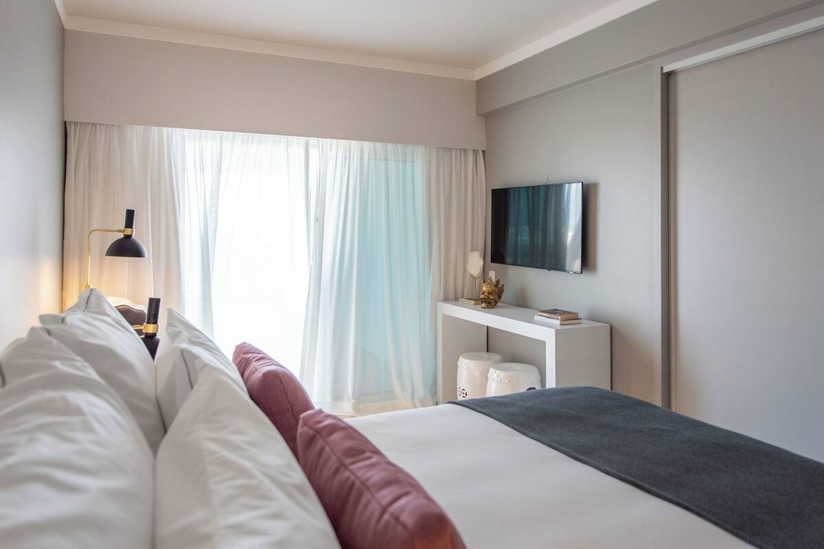 A hotel room with a bed, a bedside table with a lamp, a TV, and a window with curtains.