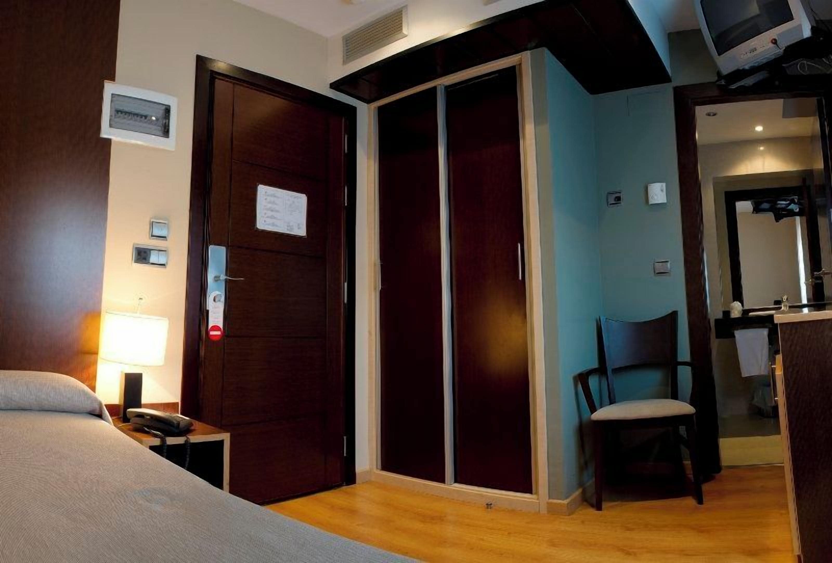A hotel room with a bed, a desk with a lamp, a chair, a wardrobe, and a television.