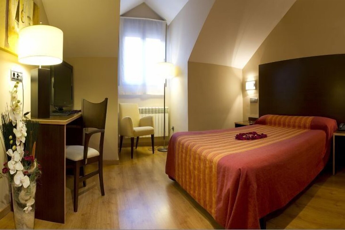 A hotel room with a bed, a desk, a chair, a lamp, and a vase with flowers.