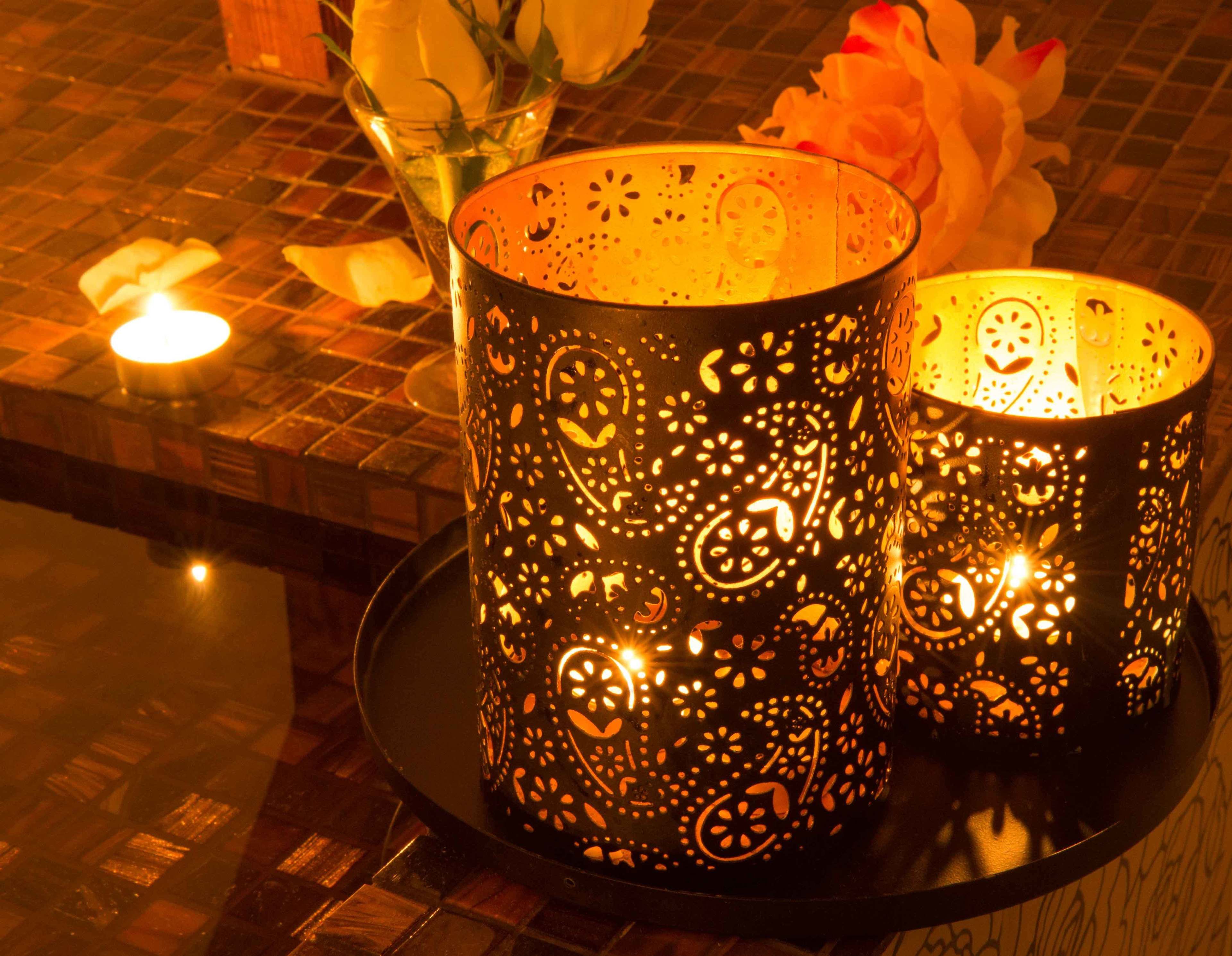 Two intricately patterned lanterns on a tray, with a candle and flowers in the background.