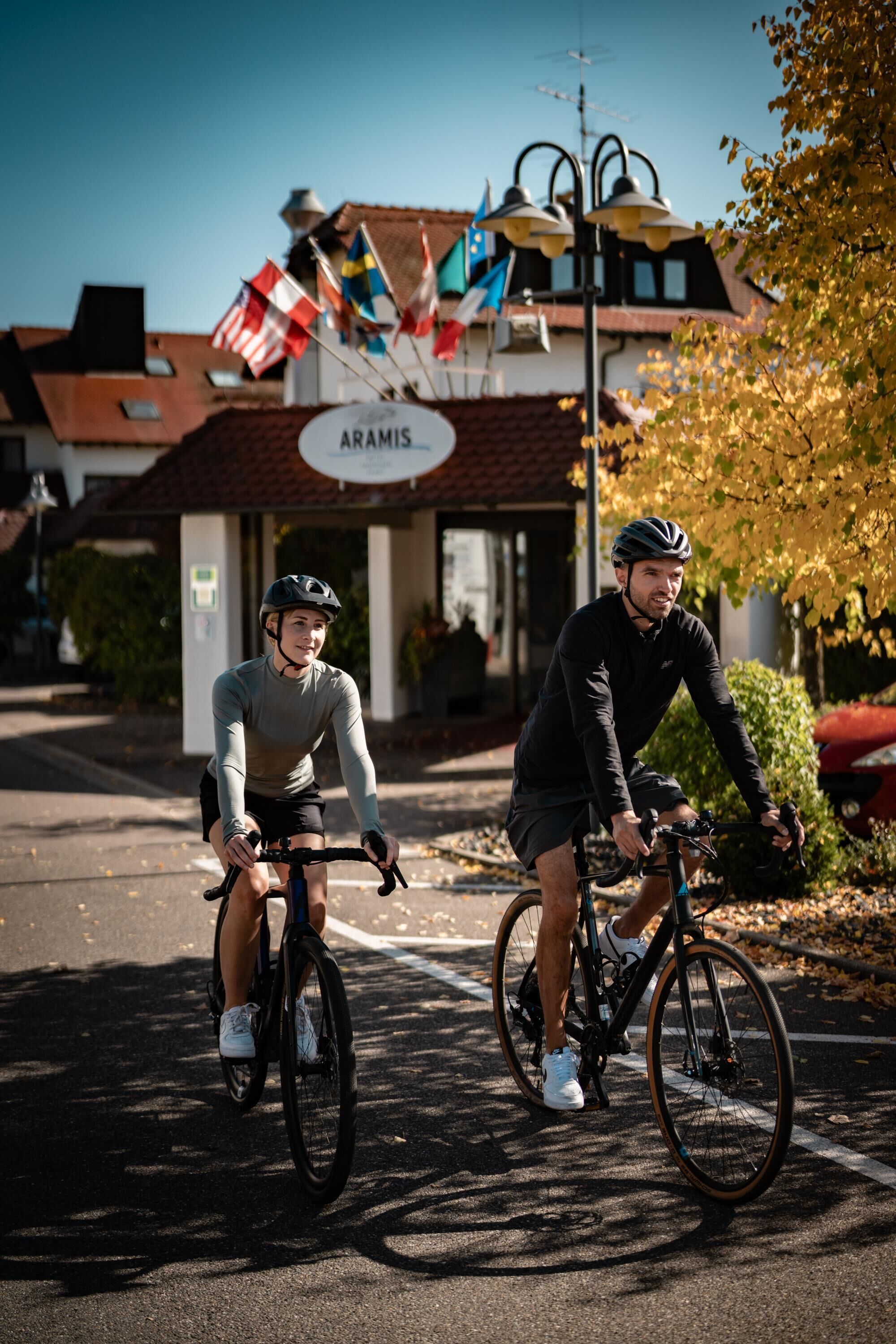 Zwei Radfahrer fahren auf einer Straße, im Hintergrund ein Gebäude mit der Aufschrift 'ARAMIS'.