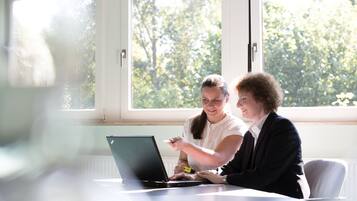 Duas pessoas sentadas à mesa com um laptop, num quarto com grandes janelas e vista para árvores.