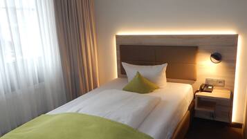 Habitación de hotel con una cama bien tendida, una mesita de noche con lámpara y una ventana con cortinas.