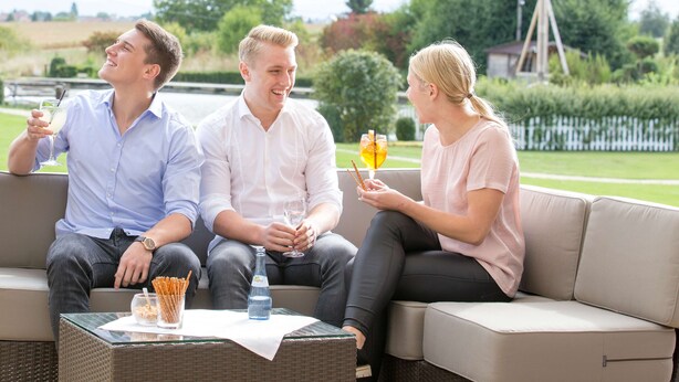 Three people sitting on a beige outdoor sofa with a table in front of them, holding drinks and glasses. - ARAMIS Tagungs- und Sporthotel (Gaeufelden)