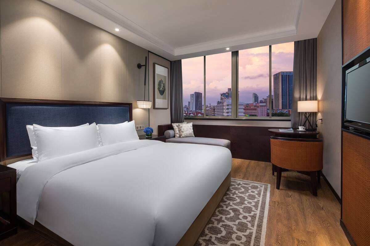 Habitación de hotel con una cama grande, un escritorio, una silla y vistas de la ciudad a través de la ventana.
