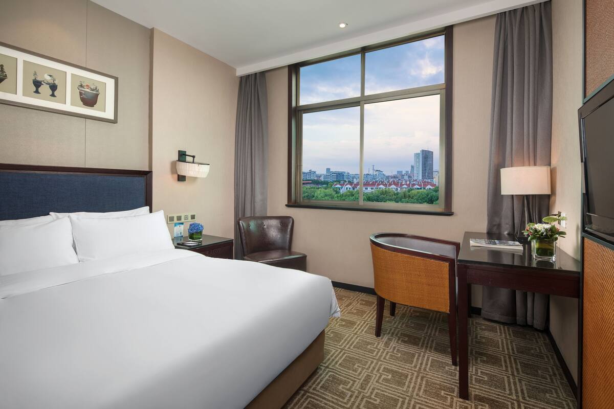 Un cuarto de hotel con una cama grande, un escritorio, una silla, vistas de la ciudad y obras de arte enmarcadas en la pared.