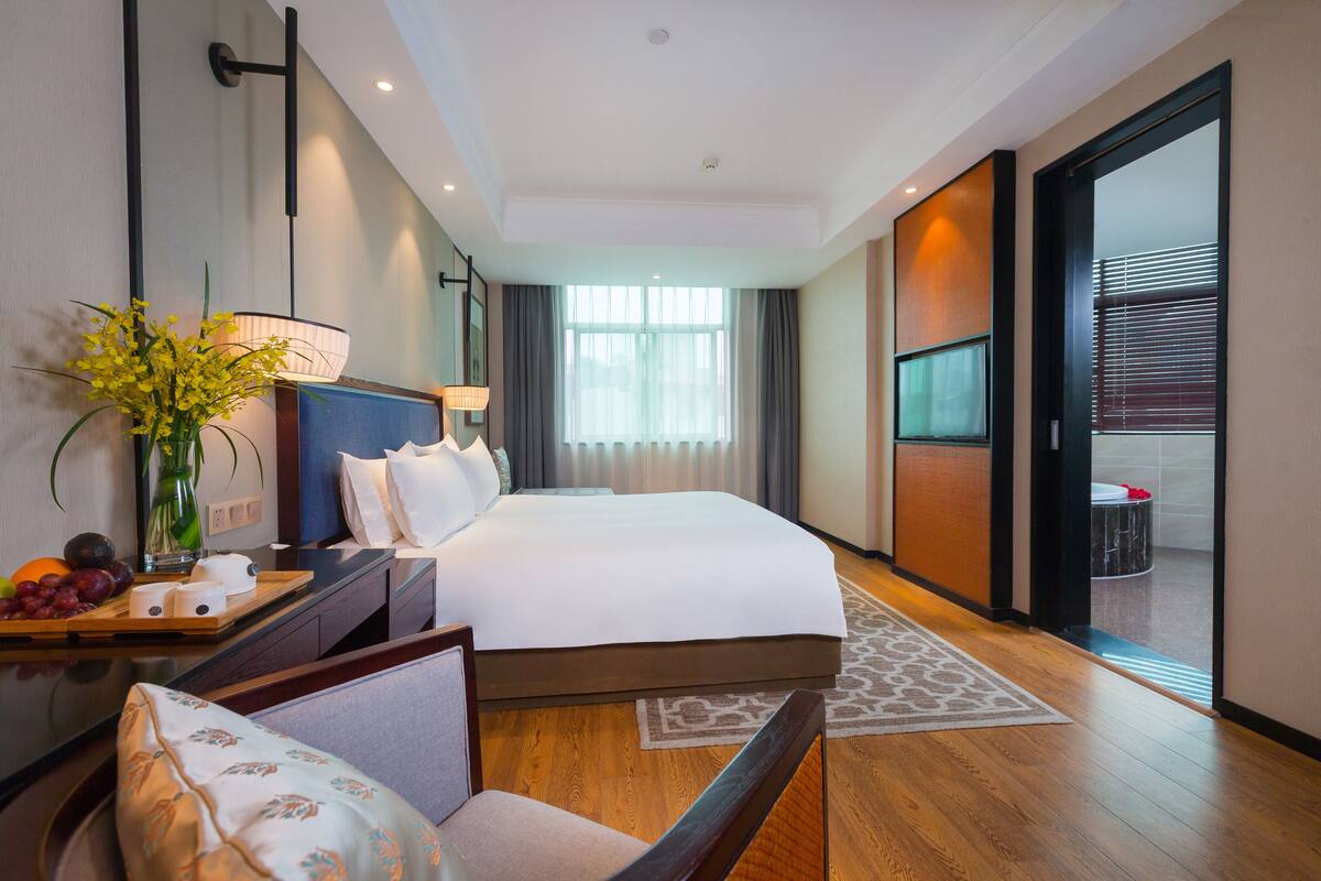 Habitación de hotel con una cama grande, mesita de noche con un jarrón de flores, televisor y vistas al exterior.
