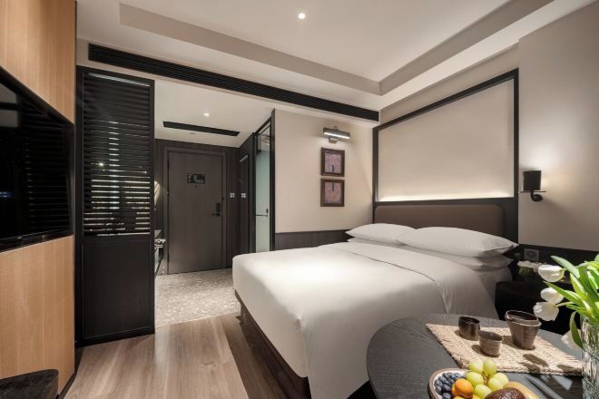 Ein modernes Hotelzimmer mit einem großen Bett, einem Nachttisch und einer kleinen Sitzecke mit einer Obstschale.