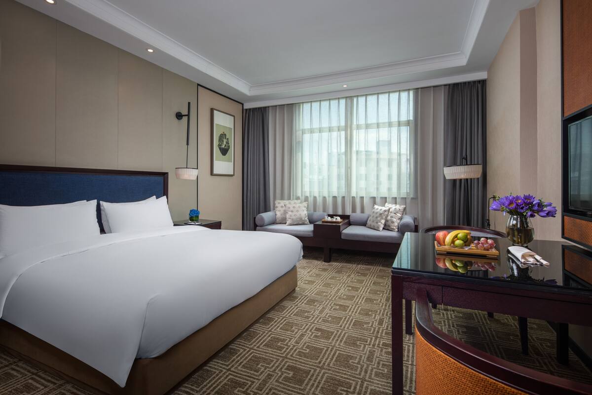 Una habitación de hotel con una cama grande, un sofá, una mesita con fruta, un televisor y una ventana con cortinas.