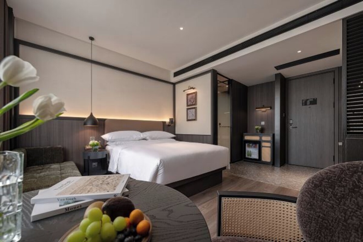 Ein modernes Hotelzimmer mit Bett, Nachttisch, Lampe und einem Essbereich mit Tisch und Stühlen.