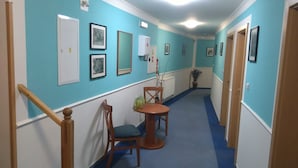 A hallway with blue walls, white trim, and a blue carpet. There are framed pictures on the walls, a wooden chair with a plant, and a small round table.