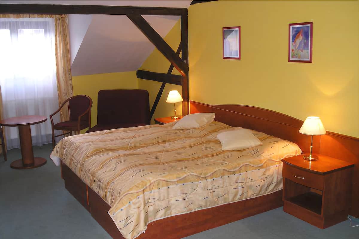 A hotel room with a bed, two bedside tables with lamps, a chair, a small table, and a window with curtains.